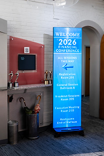 Blue portable conference banner in a hallway reading 'Welcome 2026 Financial Conference' with session list and room numbers.