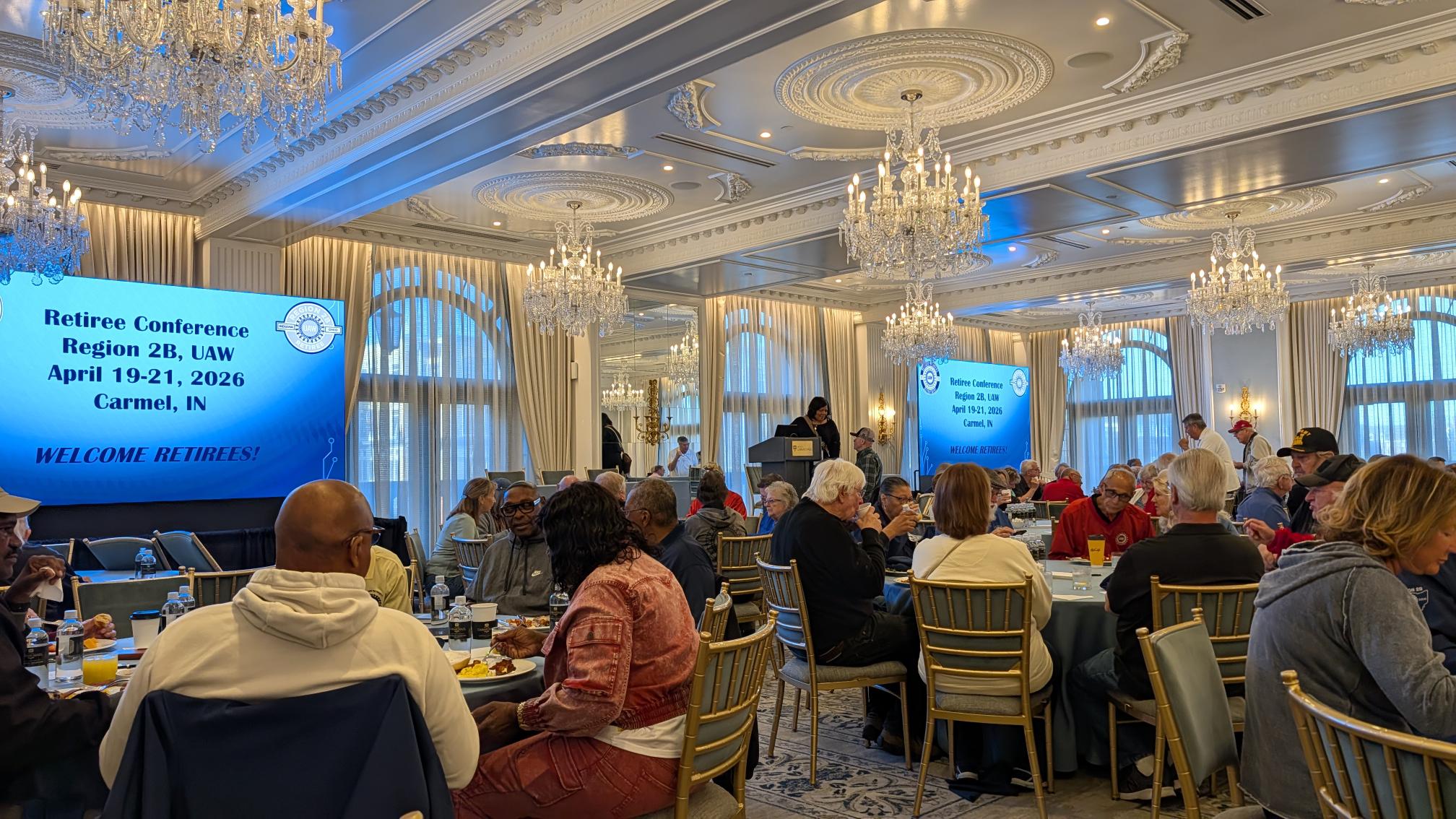 Large banquet hall with crystal chandeliers and blue conference screens reading 'Retiree Conference Region 2B, UAW, April 19-21, 2026, Carmel, IN' and 'WELCOME RETIREES!'
