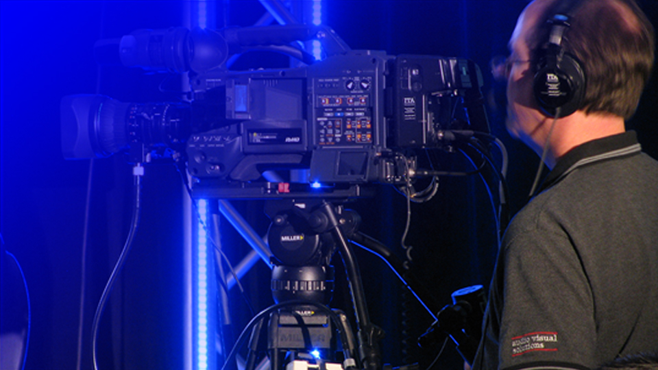 Cinematographer wearing headphones operating a professional video camera on a tripod in a blue-lit studio setting.